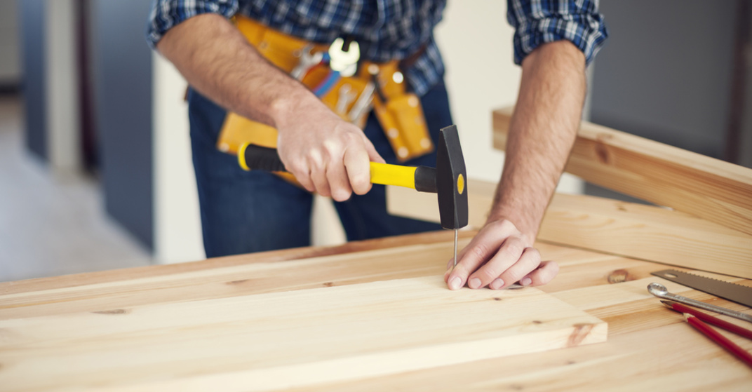 Heimwerker mit Werkzeuggürtel und in kariertem Hemd schlägt mit einem Hammer einen Nagel in ein Holzbrett. 