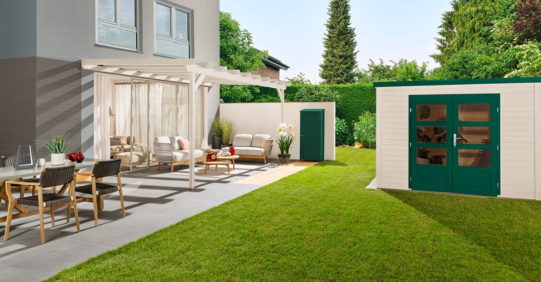 Terrasse mit Tisch und Sesseln vor Haus, gepflegter kurzer Rasen und rechts eine Gartenhütte aus Holz mit grün gestrichener Tür, blauer Himmel