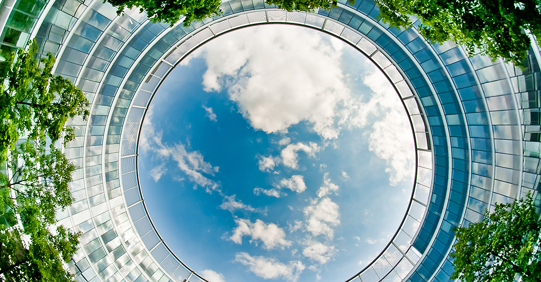 Blick vom Innenhof eines gläsernes Bürogebäudes nach oben zum wolkendurchzogenen blauen Himmel