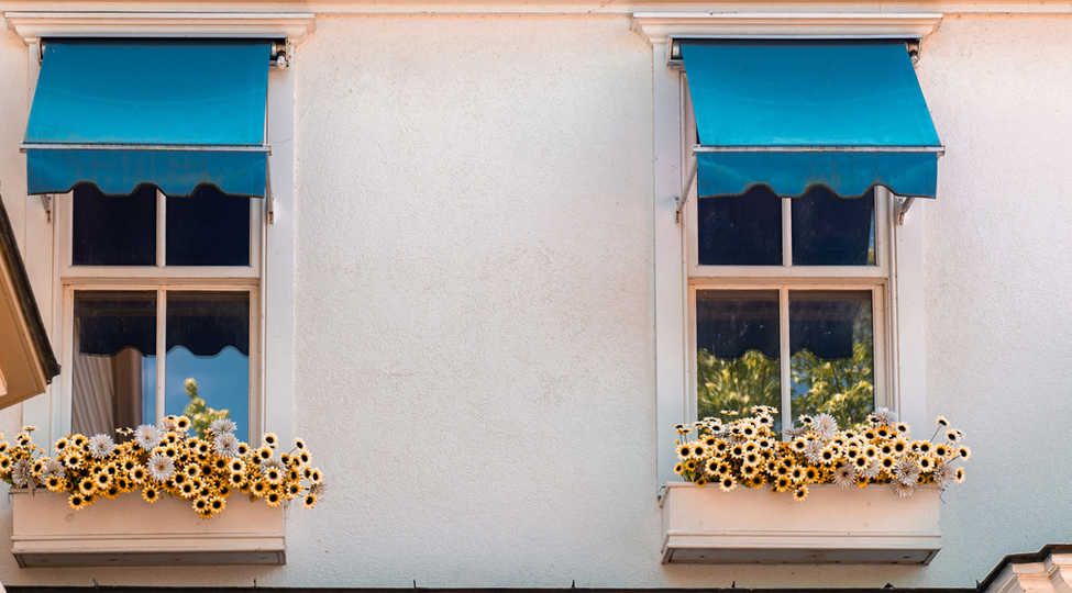 Hausfassade mit zwei kleinen Kastenfenstern, darüber tieflbaue, mediterran wirkende Fenstermarkisen. Blumen in Trögen hängen vor den Fenstern. Sonniges Ambiente